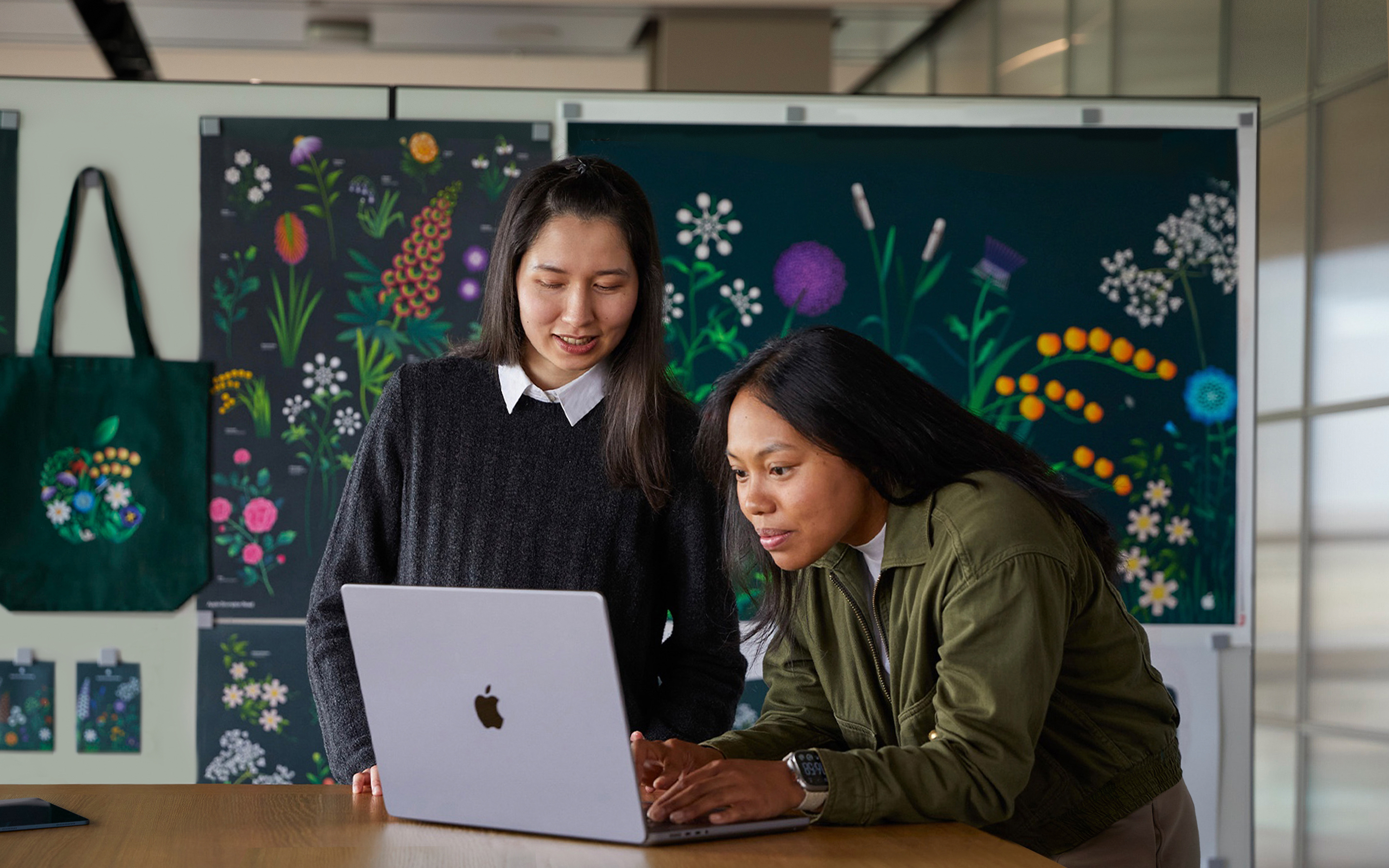 Två medarbetare i Apple Marketing-teamet som jobbar ihop på en MacBook och bakom dem syns affischer som teamet har skapat.