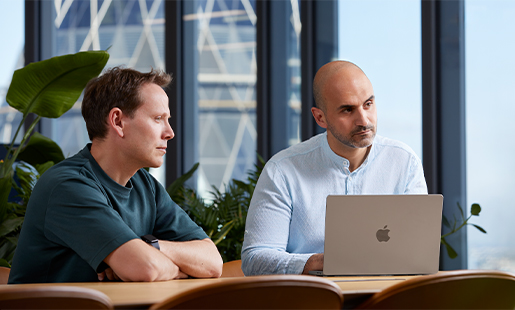 Ahmed sitter vid ett bord tillsammans med en kollega och tittar på någon utanför bild. Han har sin MacBook framför sig.