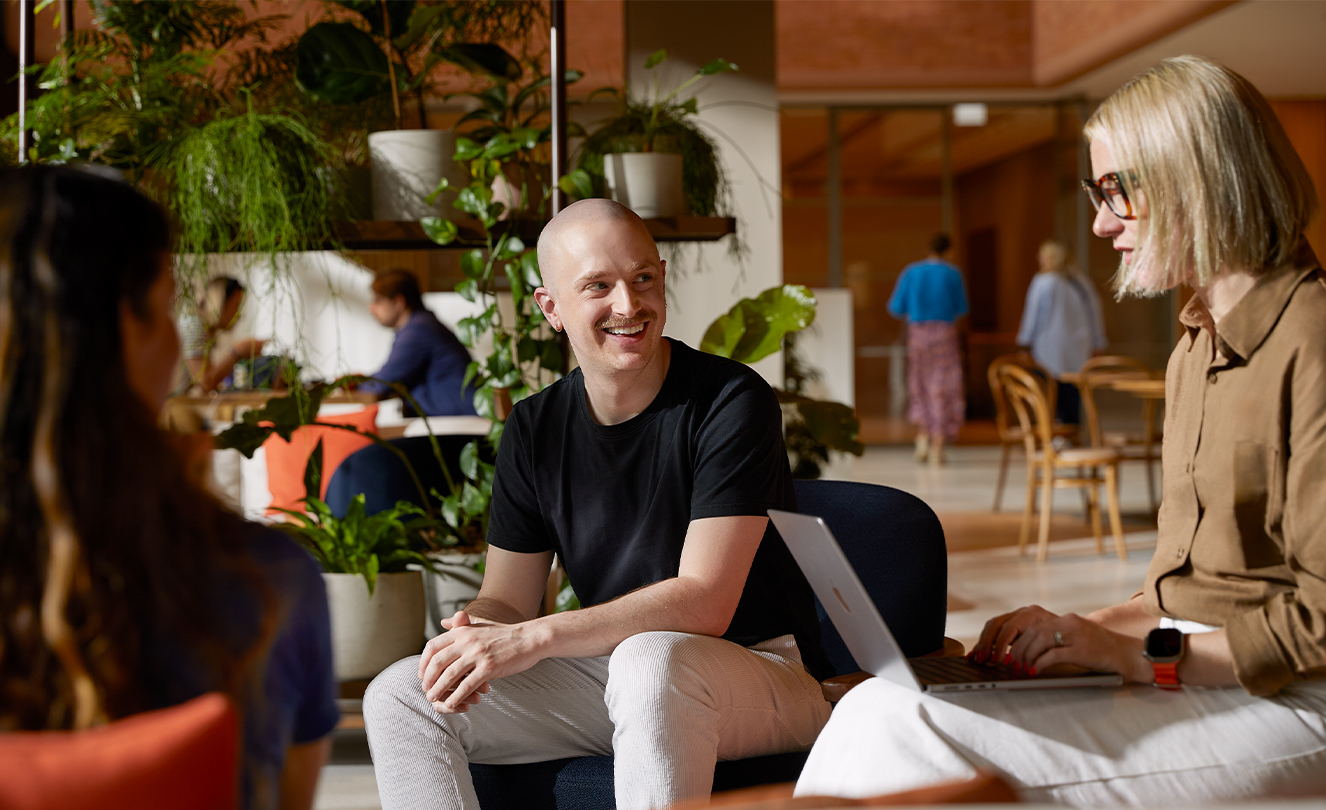 Tre Apple-medarbetare, en med en MacBook, sitter i fåtöljer i Caffè Macs och pratar med varandra.