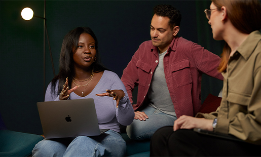Ginelle sitter med en MacBook i knät medan hon samarbetar med två kollegor.