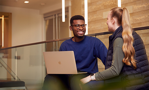 Chidi hat ein MacBook auf dem Schoss, während er mit einer Kollegin spricht. 
