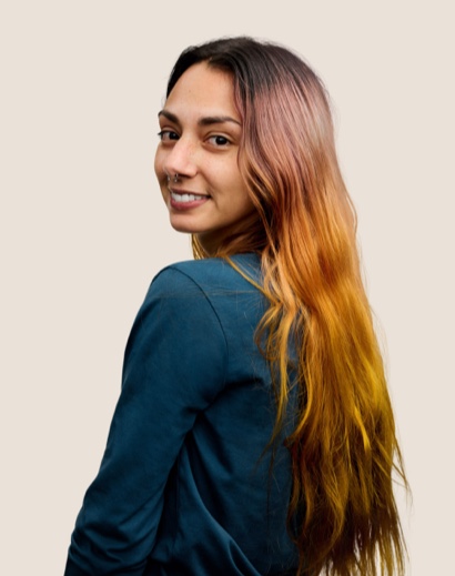 Portrait d’un membre de l’équipe Apple Retail souriant, avec de longs cheveux bruns, regardant l’objectif par-dessus son épaule.