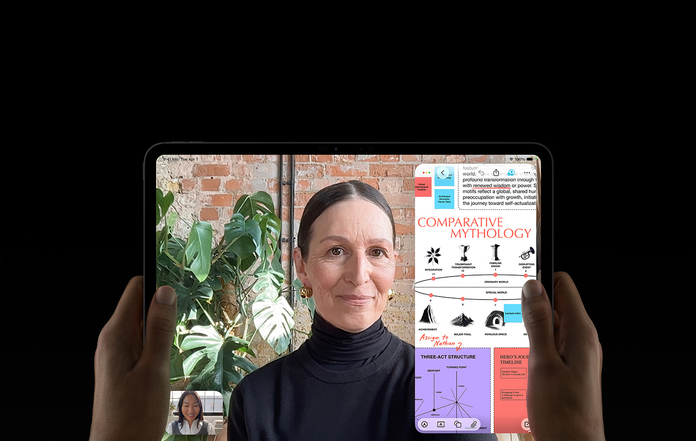 iPad Pro, front exterior, space black color, left and right hands holding in landscape orientation, rounded corners, black display bezel, display showing a FaceTime conversation being live translated, and an educational app showing a presentation of 'Comparative Mythology' with text, diagrams, and handwritten notes