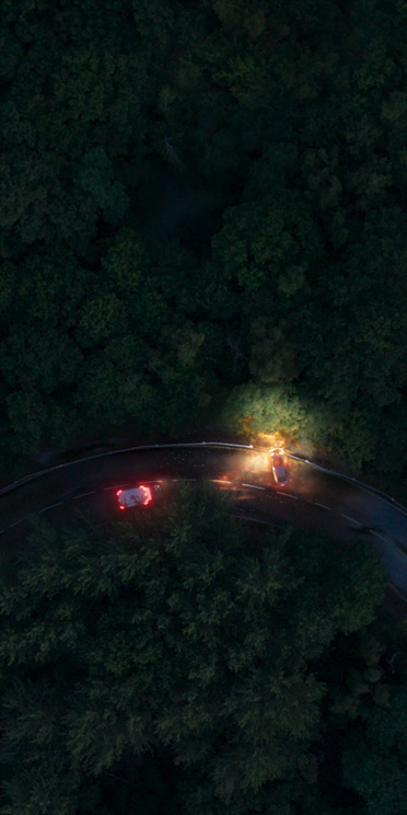 An overheard view of a winding road at night, two vehicles perpendicular with headlines on, surrounded by trees