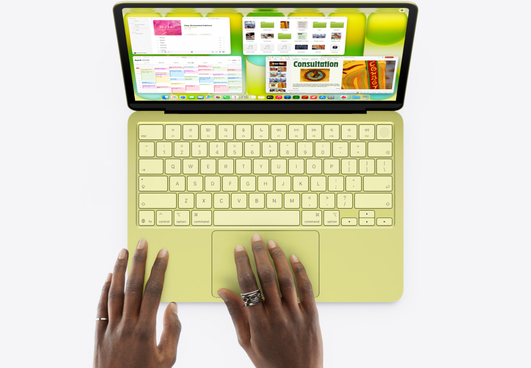 MacBook Neo цитринового кольору, рука людини на трекпеді, відкрито кілька вікон, у тому числі додатки «Музика», «Календар» та Microsoft PowerPoint