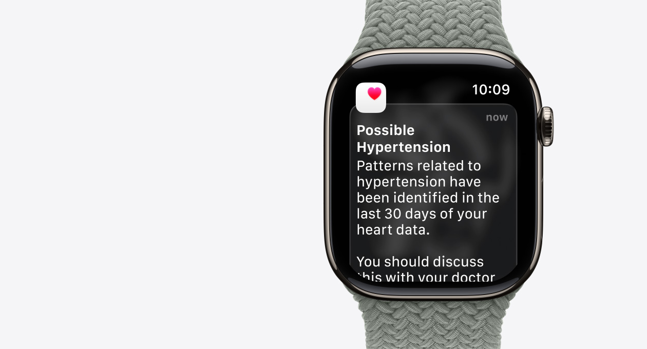 Apple Watch Series 11, prírodná farba, hlásenie o možnej hypertenzii, ružové srdce v bielom poli, pohľad zvonku na pravú stranu, korunka Digital Crown, zelenkavá šedá zapletaná slučka Solo