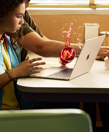 Een persoon probeert iets te pakken in een restaurant en gooit een drankje om over een geopende MacBook