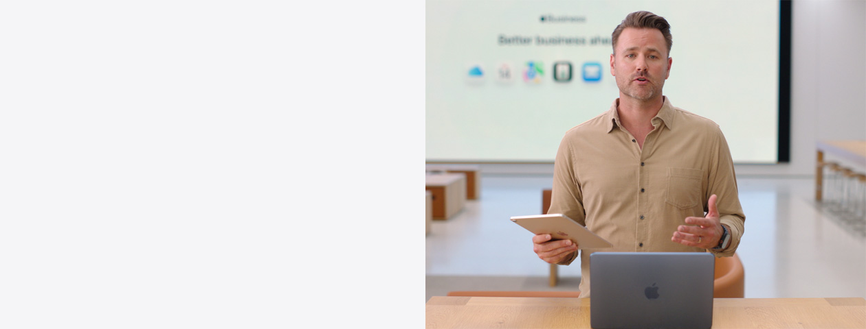 A apresentadora fala de pé, com um iPad na mão, em frente a um MacBook colocado numa mesa. Um ecrã grande apresenta vários ícones de apps empresariais.