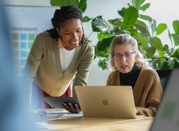 Twee docenten die samenwerken aan een tafel, de een houdt een iPad vast, de ander werkt op Mac