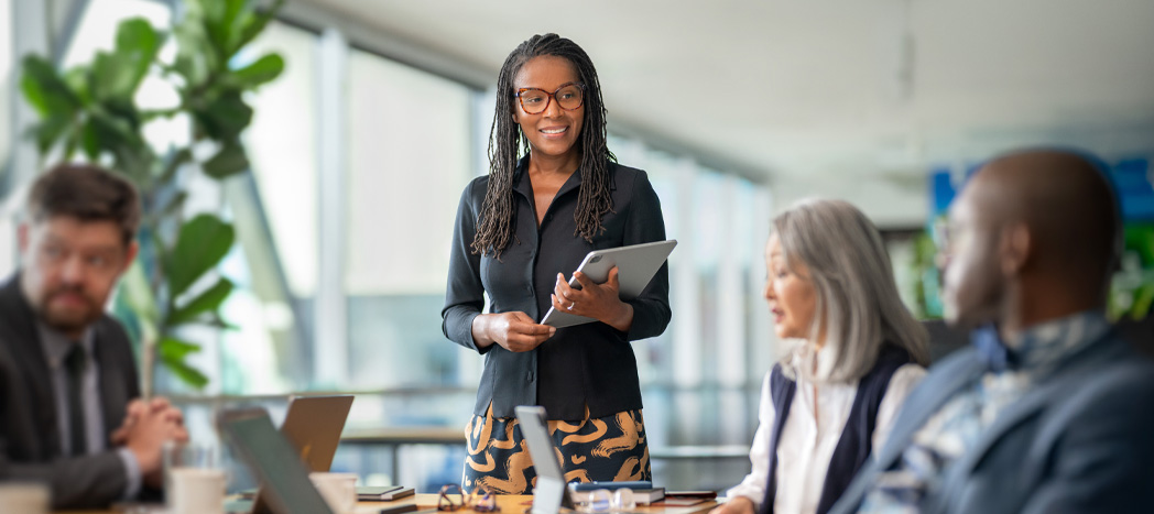 Een universitair medewerker met een iPad die een presentatie geeft, collega’s met opengeklapte MacBooks luisteren aandachtig