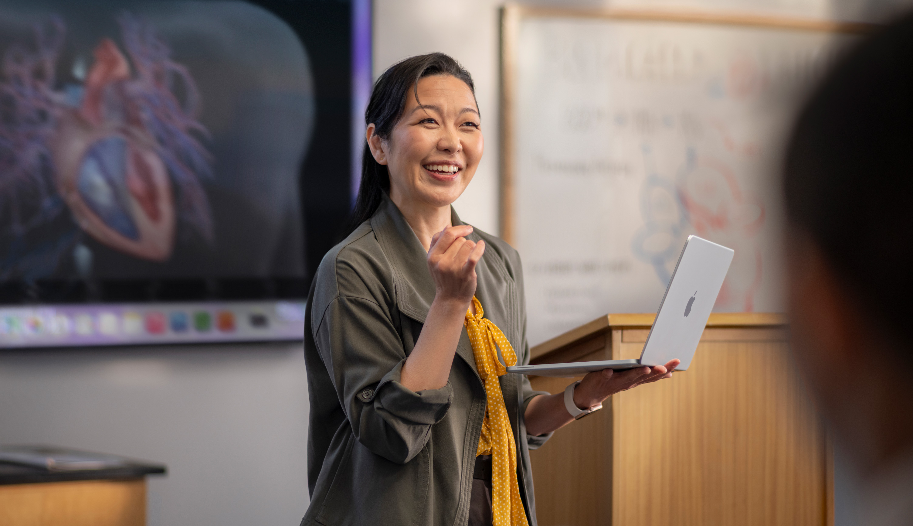 Een docent die een presentatie geeft in een collegezaal en een MacBook vasthoudt, ter illustratie van de prestaties en lange batterijduur