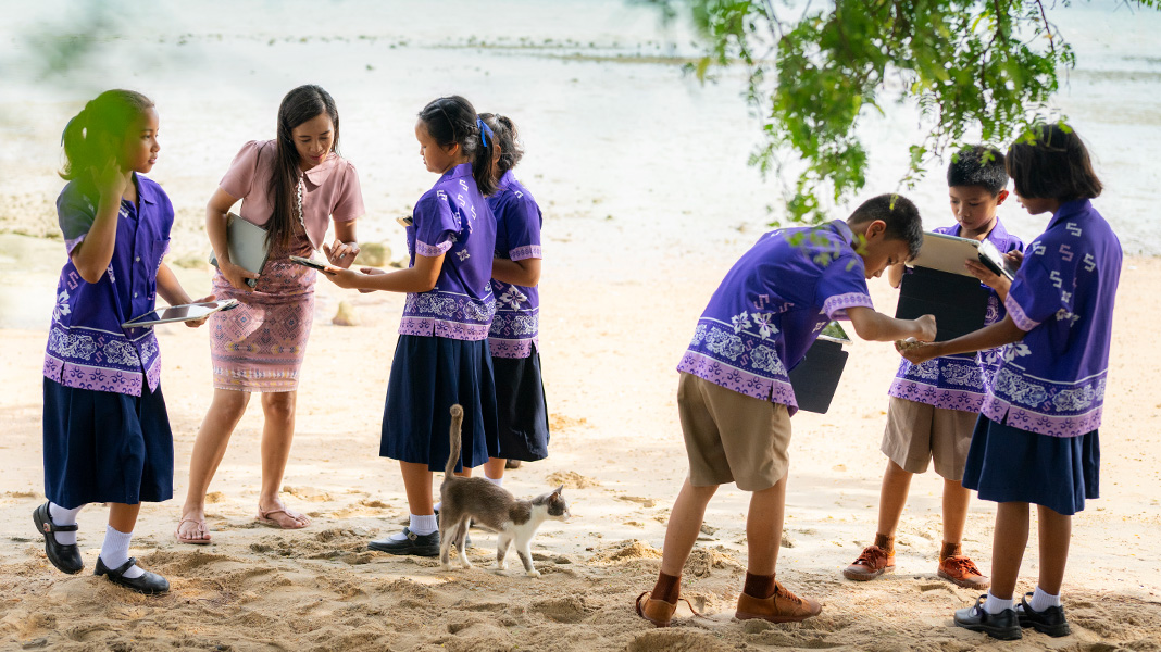 Six elementary students in colorful purple uniforms stand on a sandy beach holding iPads. A teacher is assisting one student on an iPad, and there is a cat walking amongst them.