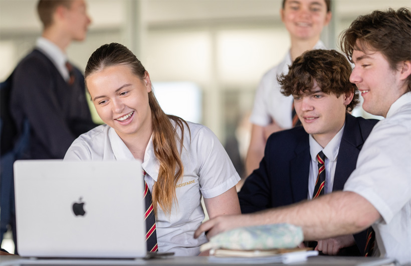 Un grupo de estudiantes con uniforme escolar se reúne alrededor de un MacBook, sonriendo y colaborando.