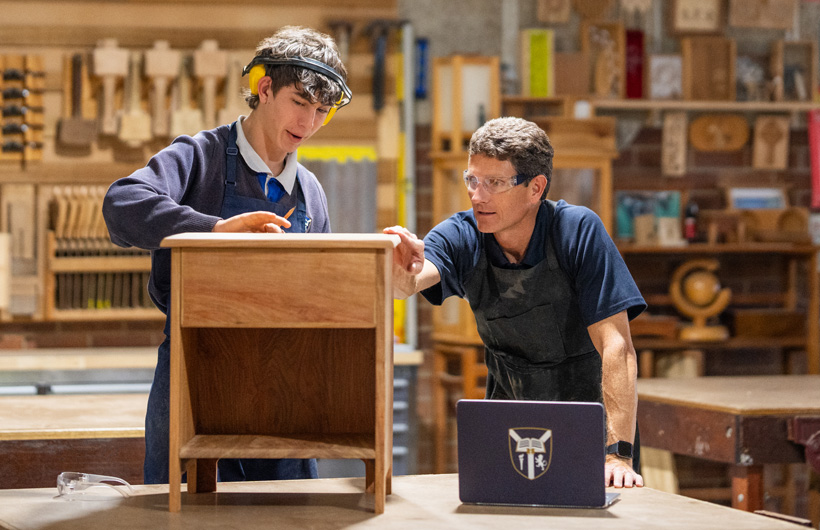 Un profesor de carpintería y un estudiante examinan un pequeño mueble de madera. Hay un portátil del colegio en la mesa de trabajo delante de ellos.