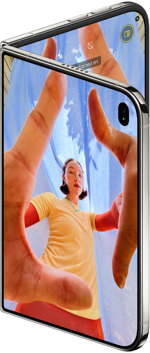 Csillagfehér iPhone Duo kissé kinyitva, a külső kijelzőn a Kamera app a Center Stage előlapi kamerával, a belső kijelzőn a Fotók app könyvtára.